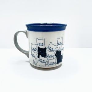 May include: A ceramic mug with a white base and a dark blue rim. The exterior features a repeating illustration of multiple cats in black and blue ink outlines. The handle is a solid light grey colour, and the mug has a smooth, cylindrical shape.