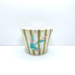 Peut inclure: Un cache-pot en céramique blanche orné de rayures verticales vertes et d'un motif floral en relief dans des tons turquoise, rose et vert. Le pot présente une forme conique cannelée avec une finition lisse et un large bord supérieur circulaire.