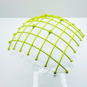 Pu&ograve; includere: Un cestino di frutta in metallo verde lime a forma di cupola. Il cestino ha un design a griglia con piccoli accenti rotondi alle intersezioni della griglia. Il cestino &egrave; su una superficie bianca.