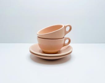 Puede incluir: Dos tazas de té de cerámica rosa apiladas con un platillo a juego. Las tazas tienen una forma redondeada y un diseño simple.