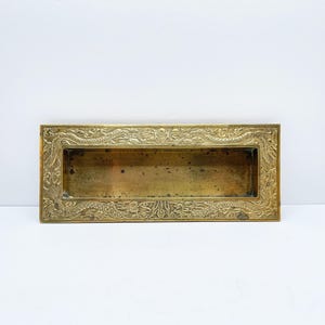 May include: A rectangular brass letterbox with an ornate, floral border. The interior is a darker, aged brass colour, suggesting age and use. The design is symmetrical and detailed.