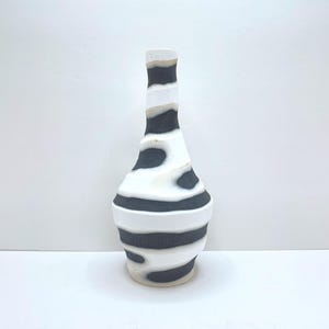 May include: A ceramic vase with a tall, slender neck and a bulbous body, featuring a distinct horizontal striped pattern in alternating matte black and smooth white bands that wrap around the entire structure from the base to the narrow opening.