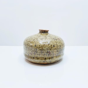 May include: A small, round ceramic vase with a speckled, textured surface in shades of olive green, brown, and red. The vase has a short, narrow neck and sits on a white surface. The overall shape is bulbous.