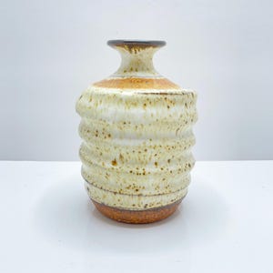 Pode incluir: Um vaso de cerâmica branco com esmalte marrom e laranja. O vaso tem um design texturizado, canelado e uma forma arredondada.