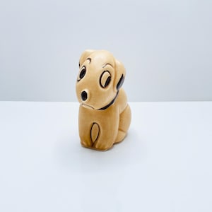 Può includere: Statuetta di cucciolo in ceramica color beige con dettagli neri. Il cane ha grandi occhi rotondi, un naso nero e un collare nero. La statuetta è seduta con le zampe ripiegate. Il cucciolo è un oggetto decorativo.