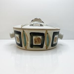 Op de afbeelding: Een crème-kleurige keramische ovenschaal met deksel. De schaal heeft een herhalend patroon van zwarte vierkanten en groene en bruine langwerpige vormen. Het deksel heeft een handvat en bijpassende patronen.