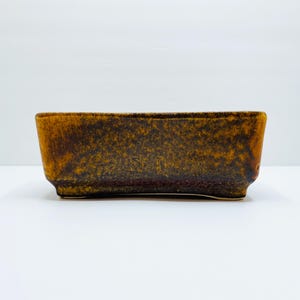 Puede incluir: Un macetero rectangular de cerámica con un esmalte marrón y amarillo. El macetero tiene un fondo ligeramente redondeado y una superficie texturizada.