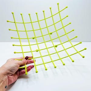 Pu&ograve; includere: Una griglia metallica verde chartreuse brillante con estremit&agrave; arrotondate. La griglia &egrave; tenuta da una mano con smalto rosso. La griglia ha una forma curva e un motivo quadrato. L'oggetto &egrave; probabilmente un oggetto decorativo o funzionale.