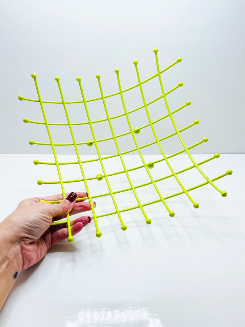 Pu&ograve; includere: Una griglia metallica verde chartreuse brillante con estremit&agrave; arrotondate. La griglia &egrave; tenuta da una mano con smalto rosso. La griglia ha una forma curva e un motivo quadrato. L'oggetto &egrave; probabilmente un oggetto decorativo o funzionale.