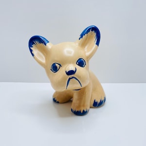 May include: A ceramic French Bulldog figurine. The dog is tan with blue detailing on the ears, eyes, nose, and paws. The figurine is in a sitting pose and has a slightly melancholic expression.