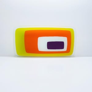 Puede incluir: Una pieza decorativa rectangular de vidrio con un diseño en capas. La capa exterior es amarilla, seguida de naranja, blanco y un centro púrpura. La pieza tiene un borde transparente y un acabado brillante.