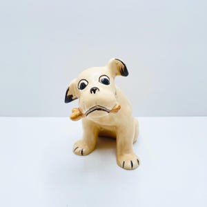 May include: A ceramic dog figurine, seated and holding a bone in its mouth. The dog is beige with black detailing on its ears and paws. The bone is tan. The figurine has a glossy finish and is set against a white background.