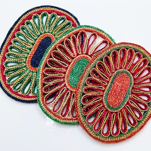 Op de afbeelding: Drie kleurrijke geweven onderzetters. Elke onderzetter heeft een centraal ovaal ontwerp omgeven door stralende lussen. De onderzetters zijn in tinten rood, groen, geel en blauw, met een textuur.