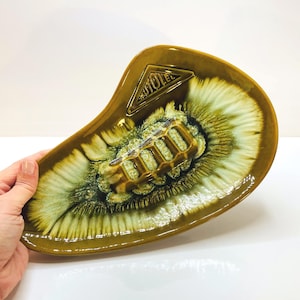 Può includere: Un vassoio in ceramica asimmetrico caratterizzato da un motivo a smalto colato lucido color verde oliva e crema. Il centro presenta una sezione rettangolare rialzata e scanalata, con un logo triangolare in rilievo che riporta AUTOLEC.