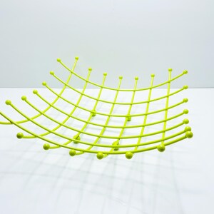 Pu&ograve; includere: Una moderna fruttiera in metallo verde lime. La ciotola ha un design curvo a griglia con accenti di palline arrotondate alle estremit&agrave; delle barre curve. La ciotola &egrave; vuota e poggia su una superficie bianca.