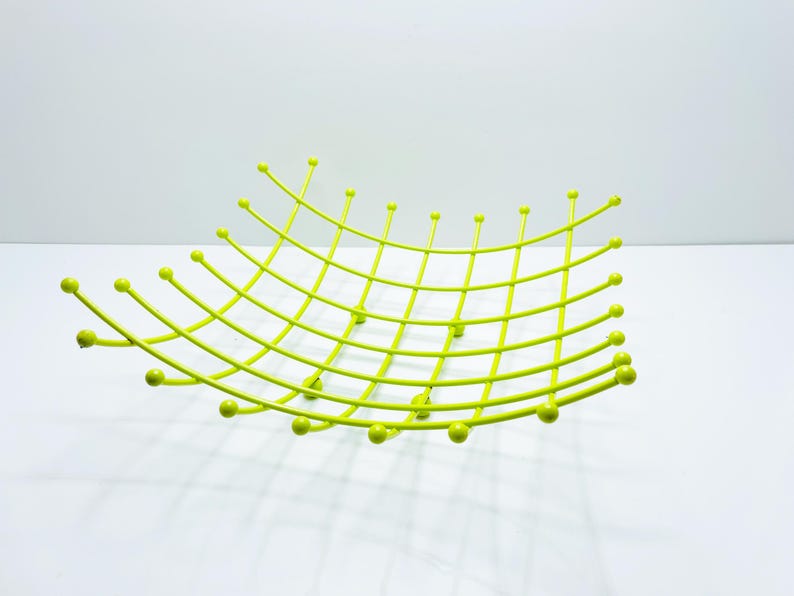 Pu&ograve; includere: Una moderna fruttiera in metallo verde lime. La ciotola ha un design curvo a griglia con accenti di palline arrotondate alle estremit&agrave; delle barre curve. La ciotola &egrave; vuota e poggia su una superficie bianca.