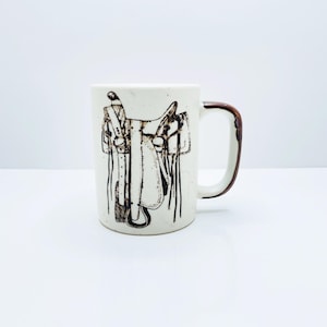 Peut inclure: Mug en céramique blanche avec une illustration détaillée d'une selle de cheval dans des tons bruns. La tasse a une finition mouchetée et une anse marron. Cette tasse sur le thème du western est idéale pour le thé ou le café.