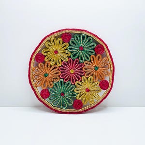 Puede incluir: Un posavasos redondo y tejido con un diseño floral. El posavasos presenta un borde rojo con ribete dorado, y el interior está lleno de flores coloridas en tonos amarillos, naranjas, verdes y rojos. Aproximadamente 15 cm de diámetro.
