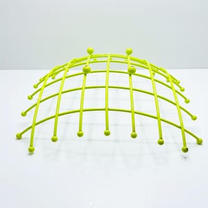 Pu&ograve; includere: Una fruttiera in filo metallico verde lime a forma di cupola. La fruttiera ha un design a griglia con barre verticali e piccoli accenti sferici alle estremit&agrave; delle barre. La fruttiera &egrave; su una superficie bianca.