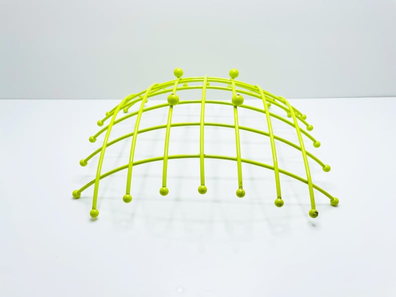 Pu&ograve; includere: Una fruttiera in filo metallico verde lime a forma di cupola. La fruttiera ha un design a griglia con barre verticali e piccoli accenti sferici alle estremit&agrave; delle barre. La fruttiera &egrave; su una superficie bianca.