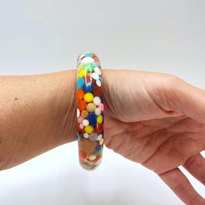 Może przedstawiać: Bransoletka typu bangle z przezroczystej żywicy z gęstym układem zatopionych, wielokolorowych, okrągłych elementów konfetti w odcieniach czerwieni, błękitu, żółci, zieleni, bieli i brązu, tworzących żywy i teksturowany wygląd na całej powierzchni.