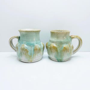 May include: Two ceramic mugs with a light green glaze and brown accents. The mugs have a rounded shape and a handle.