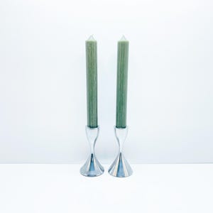 Può includere: Due candele affusolate verdi in candelabri in metallo argentato. I candelabri hanno una base svasata e uno stelo curvo a forma di clessidra. Le candele sono di un verde tenue e sono centrate su uno sfondo bianco.