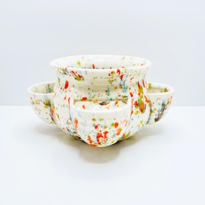 May include: A white ceramic planter with a speckled design of red, orange, green, and blue paint splatters. The planter has a rounded shape with three smaller, attached cup-like compartments. A unique, decorative piece for plants.