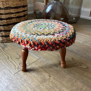 May include: A round footstool featuring a thick, multi-coloured braided fabric top with concentric patterns in shades of red, blue, white, and brown. The stool is supported by three short, smooth, dark brown wooden legs with a turned design.