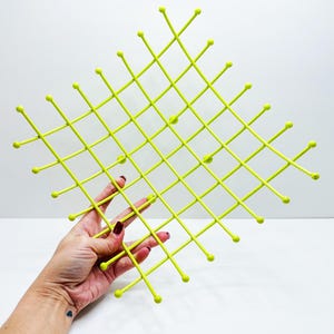Pu&ograve; includere: Un elemento decorativo da parete, di colore verde chartreuse brillante, con un motivo a griglia. La griglia &egrave; composta da linee sottili e dritte con piccoli pomelli arrotondati alle intersezioni e alle estremit&agrave;. L'oggetto &egrave; tenuto da una mano.