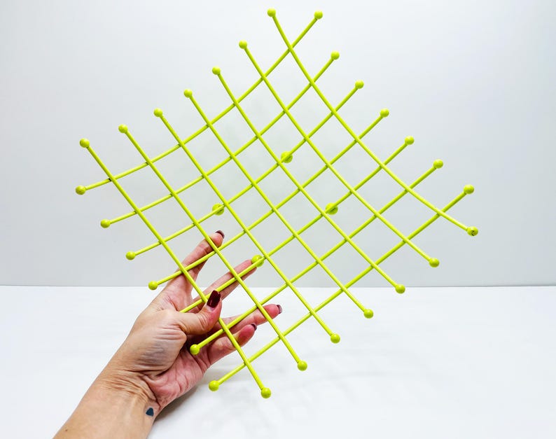 Pu&ograve; includere: Un elemento decorativo da parete, di colore verde chartreuse brillante, con un motivo a griglia. La griglia &egrave; composta da linee sottili e dritte con piccoli pomelli arrotondati alle intersezioni e alle estremit&agrave;. L'oggetto &egrave; tenuto da una mano.