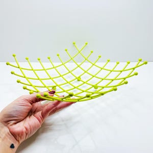 Pu&ograve; includere: Un cestino di frutta in filo metallico color chartreuse con un design a griglia. La ciotola ha una forma poco profonda e aperta con estremit&agrave; arrotondate e piccoli accenti a sfera alle intersezioni del filo. La ciotola &egrave; tenuta da una mano su sfondo bianco.