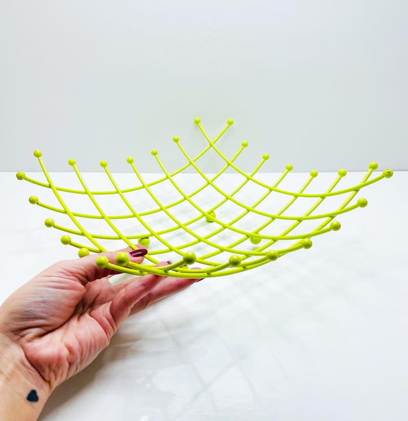 Pu&ograve; includere: Un cestino di frutta in filo metallico color chartreuse con un design a griglia. La ciotola ha una forma poco profonda e aperta con estremit&agrave; arrotondate e piccoli accenti a sfera alle intersezioni del filo. La ciotola &egrave; tenuta da una mano su sfondo bianco.