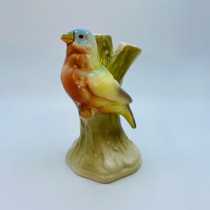 Puede incluir: Una figurita de pájaro de cerámica posada en un tronco de árbol, pintada en tonos de amarillo, verde, azul y marrón. El pájaro tiene un pico amarillo y detalles azules en la cabeza. El tronco del árbol es verde con detalles marrones. Esta figurita es una pieza decorativa para un hogar o jardín.