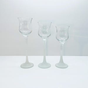 Könnte beinhalten: Drei klare Glaskerzenhalter mit einer tulpenförmigen Schale und einem langen, schlanken Stiel. Die Sockel sind mattiert und die Kerzenhalter sind auf einer weißen Oberfläche angeordnet. Die Kerzenhalter sind unterschiedlich hoch.
