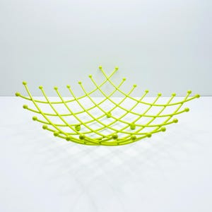 Pu&ograve; includere: Un cestino di frutta in metallo verde lime brillante con un design geometrico aperto. Il cestino presenta un motivo a forma di diamante con piccoli accenti arrotondati. Il cestino &egrave; su una superficie bianca.