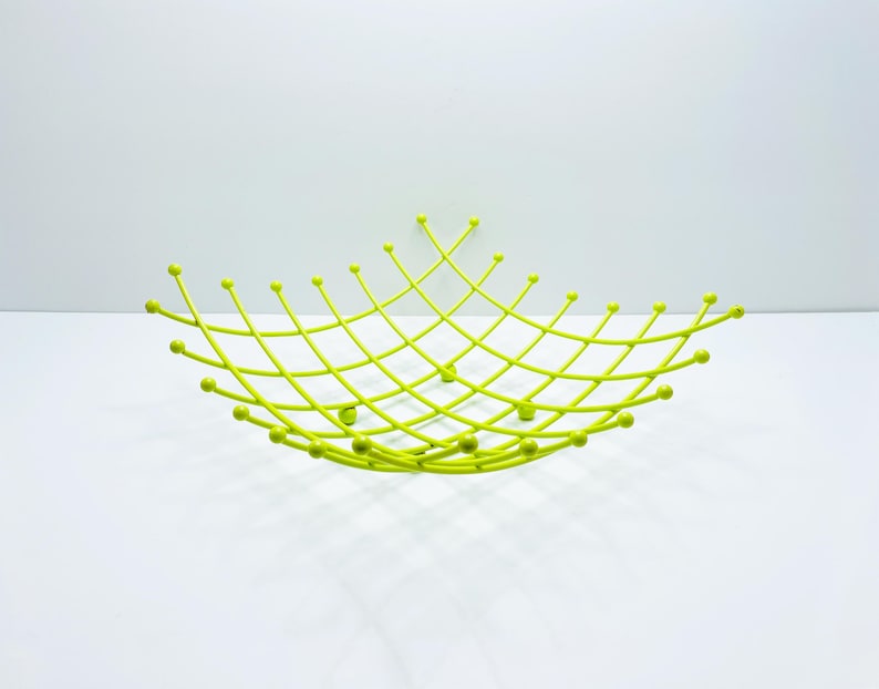 Pu&ograve; includere: Un cestino di frutta in metallo verde lime brillante con un design geometrico aperto. Il cestino presenta un motivo a forma di diamante con piccoli accenti arrotondati. Il cestino &egrave; su una superficie bianca.