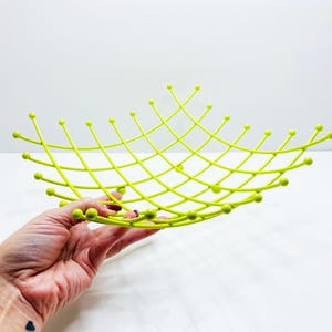 Pu&ograve; includere: Una fruttiera in metallo traforato color chartreuse. La ciotola ha un design a griglia con punte arrotondate alle intersezioni. La ciotola &egrave; tenuta in mano da una persona su sfondo bianco. La ciotola &egrave; larga circa 30 cm.