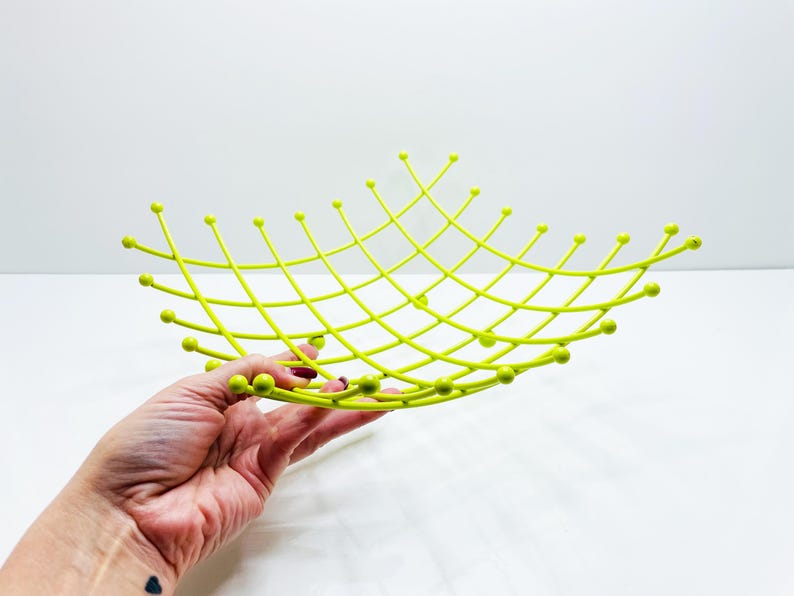 Pu&ograve; includere: Una fruttiera in metallo traforato color chartreuse. La ciotola ha un design a griglia con punte arrotondate alle intersezioni. La ciotola &egrave; tenuta in mano da una persona su sfondo bianco. La ciotola &egrave; larga circa 30 cm.