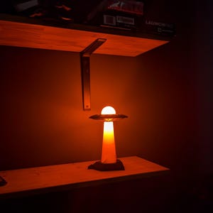 May include: A unique UFO-shaped lamp with a bright, glowing orb at the top. The lamp has a cylindrical base and a saucer-like top, emitting a warm, orange light. It sits on a wooden shelf, creating a sci-fi ambiance.