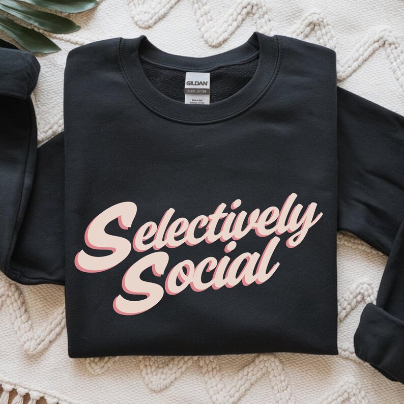 A folded black sweatshirt with the words "Selectively Social" written in a light pink script.