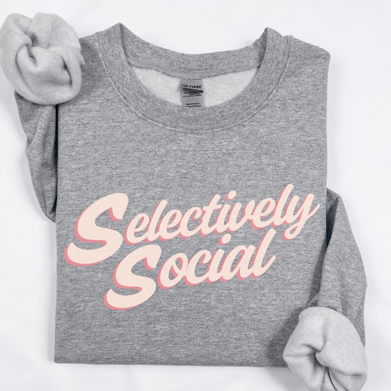 A folded gray sweatshirt with the words "Selectively Social" written in a light pink script.