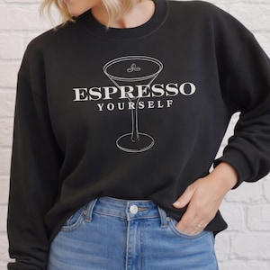 May include: A black long-sleeve crewneck sweatshirt featuring a white line-art graphic of a martini glass containing three coffee beans. The text ESPRESSO YOURSELF is printed in white capital letters across the center of the graphic design.
