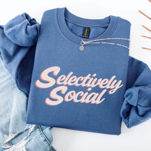 A folded blue sweatshirt with the words "Selectively Social" written in a light pink script.