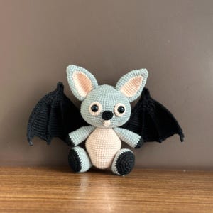 May include: A crocheted bat plush toy with black wings, light blue body, pink ears, and black eyes.