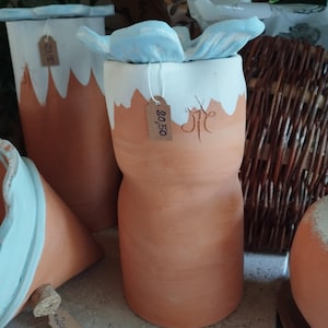 Op de afbeelding: Terracotta-kleurige keramische vaas met een witte, golvende bovenkant en een lichtblauw decoratief element. De vaas heeft een unieke zandloper vorm. Een prijskaartje is bevestigd. Andere soortgelijke vazen staan op de achtergrond.