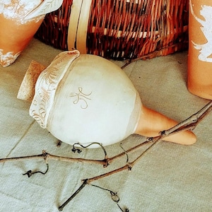 Può includere: Una bottiglia di ceramica bianca con un tappo di sughero e un design decorativo. La bottiglia ha un beccuccio lungo e sottile e poggia su un ramo.