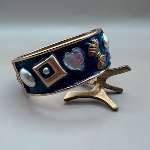 May include: A gold bangle bracelet with a blue enamel base and decorative accents. The accents include a heart, a bow, and a square with a pearl in the center. The bracelet is on a gold stand.