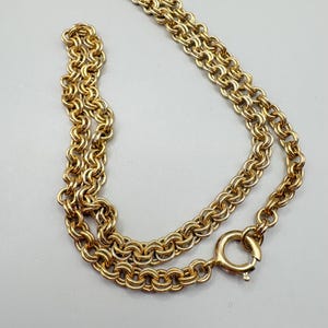 May include: A gold-colored necklace with a double-strand chain design. The chain is made of interlocking circular links, and it has a round clasp. The necklace is displayed on a light gray surface.