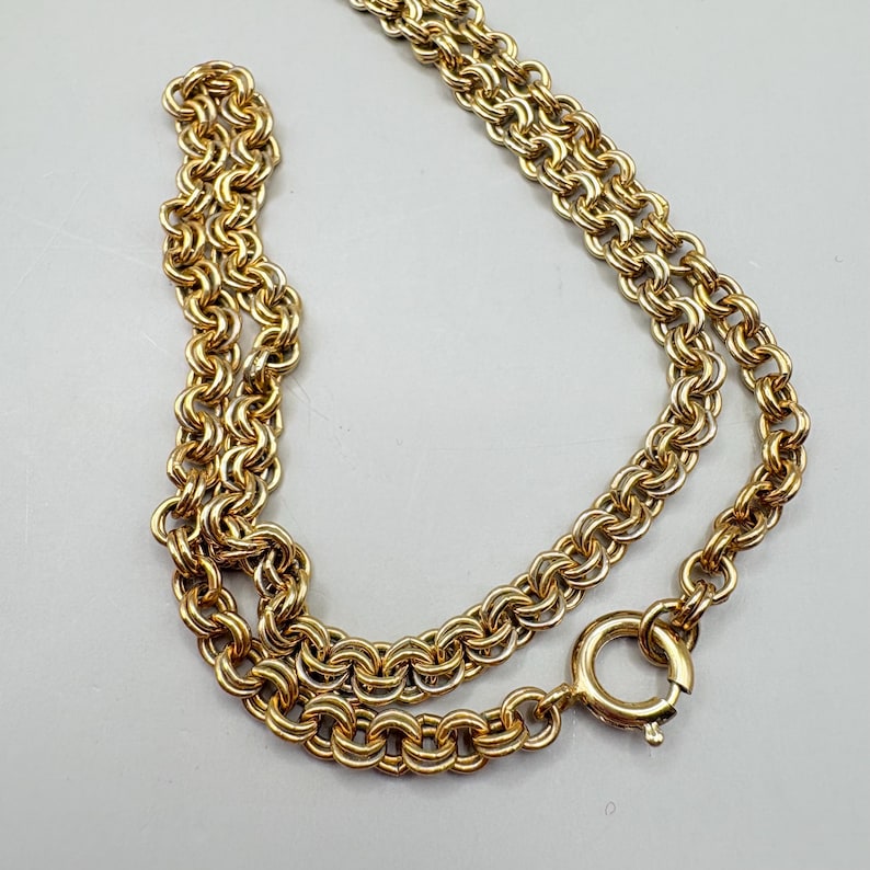 May include: A gold-colored necklace with a double-strand chain design. The chain is made of interlocking circular links, and it has a round clasp. The necklace is displayed on a light gray surface.
